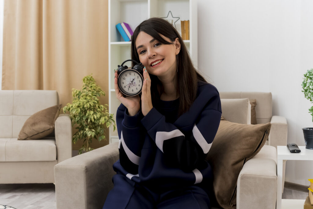 Smiling Young Pretty Caucasian Girl Sitting On Armchair In Designed Living Room Holding Alarm Clock Looking At Camera - RW Office