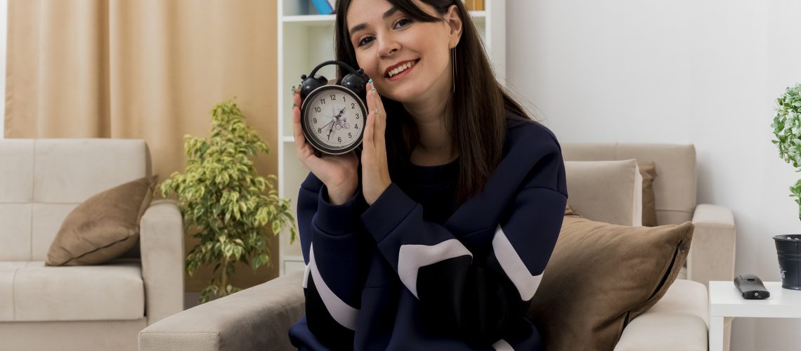 Smiling Young Pretty Caucasian Girl Sitting On Armchair In Designed Living Room Holding Alarm Clock Looking At Camera - RW Office