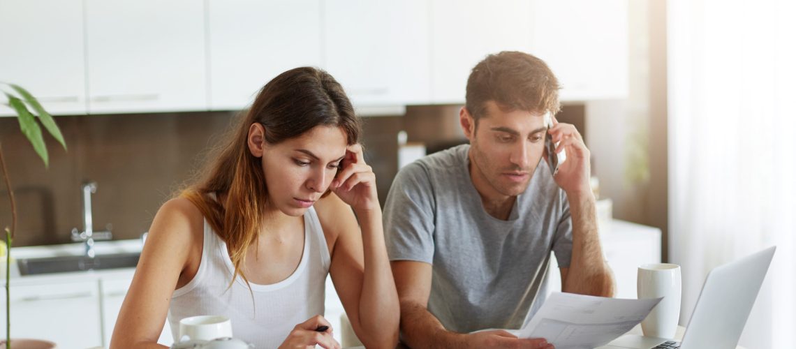 Indoor Shot Of Serious Couple Of Business Workers Being Busy At Home While Reading Attentively Documents, Making Financial Report And Calculating Annual Figures, Making Arrangements Over Mobile Phone - RW Office